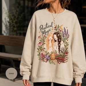 Peut inclure: Un sweat-shirt à col rond couleur sable avec une impression graphique représentant deux femmes entourées de fleurs, un croissant de lune, un bocal étiqueté Rose Petals, une bougie, un mortier et un pilon, et des livres étiquetés Herbs, Spells, Remedies.