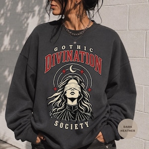 May include: A dark heather gray crewneck sweatshirt featuring a graphic print with the text GOTHIC DIVINATION SOCIETY. The design includes a central illustration of a person with a blindfold, a crescent moon, and stars in red and white ink.