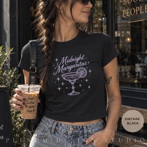 Può includere: Una t-shirt nera a maniche corte con una stampa grafica bianca di un bicchiere da margarita con una fetta di lime, circondata da icone a forma di stella e dal testo Midnight Margaritas. La maglietta ha un girocollo e un orlo corto.