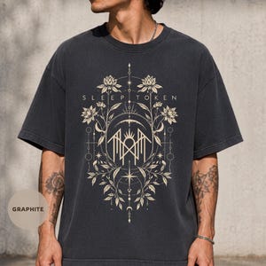 May include: A graphite-colored short-sleeve t-shirt featuring a cream-colored graphic print. The design includes the text "SLEEP TOKEN" above a central geometric emblem surrounded by intricate floral motifs, stars, and celestial line art elements.