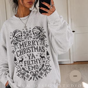 Può includere: Una felpa girocollo grigio chiaro con una stampa grafica nera che riporta il testo MERRY CHRISTMAS YA FILTHY EMO. Il design include teschi con cappelli da Babbo Natale, fiocchi di neve, renne, agrifoglio e bastoncini di zucchero.