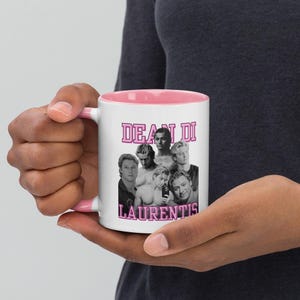 Könnte beinhalten: Ein weißer Keramikbecher mit rosa Innenseite und Henkel, der eine schwarz-weiße Fotocollage eines Mannes in verschiedenen Posen zeigt. Der Text DEAN DI LAURENTIS ist in fetter rosa Schrift über und unter der Collage auf der Vorderseite gedruckt.