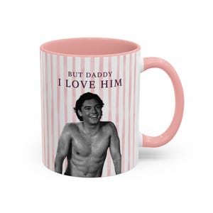 May include: A ceramic mug with a pink interior and handle, featuring a white and pink vertical striped background. The mug displays a black and white photograph of a shirtless person and the text BUT DADDY I LOVE HIM in dark purple lettering.