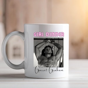 Puede incluir: Una taza de cerámica blanca con el texto rosa GIRL DINNER en la parte superior y una fotografía en blanco y negro de una persona sin camisa haciendo ejercicio en una barra de dominadas en el centro, con el texto cursivo negro Garret Graham debajo.