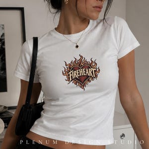 Op de afbeelding: Een wit T-shirt met korte mouwen en een centrale afbeelding van een donkerrood hart omringd door gestileerde oranje en gele vlammen. Het woord FIREHEART is in een vet, decoratief lettertype over het midden van het hart gedrukt.