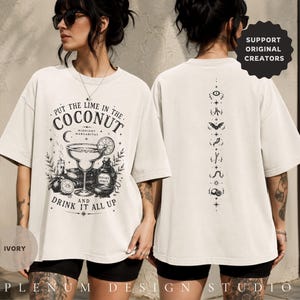 May include: An ivory short-sleeve t-shirt featuring black graphic prints. The front displays a margarita glass, lime, and text reading "PUT THE LIME IN THE COCONUT" and "DRINK IT ALL UP." The back features a vertical column of small mystical symbols.