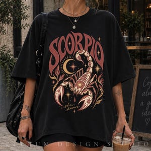 以下が含まれることがあります： 赤と金のグラフィックプリントが施された黒の半袖Tシャツ。サソリ、三日月、星が描かれています。サソリの上にはSCORPIOの文字がアーチ状に配置され、サソリの下にはshe knowsという文字がプリントされています。