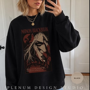 May include: A black crewneck sweatshirt featuring a graphic print of a character with long hair and a dragon silhouette. The text reads MANON BLACKBEAK and From now until the darkness claims us. The design uses dark red and cream tones on the black fabric.
