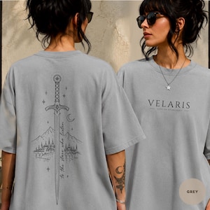May include: A grey short-sleeve t-shirt featuring a black line art graphic on the back of a sword, mountains, a city, and the text "to the stars who listen." The front of the shirt displays the text "VELARIS CITY OF STARLIGHT" in black lettering.