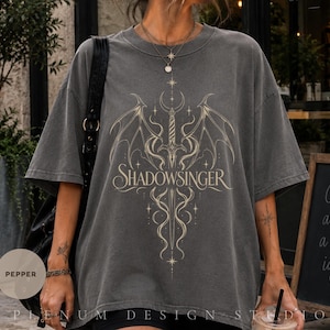 Könnte beinhalten: Ein anthrazitfarbenes Kurzarm-T-Shirt mit einem hellen Grafikdruck, der ein Schwert mit Flügeln und den Schriftzug "SHADOWSINGER" mittig auf der Brust zeigt. Das Design enthält sternförmige Akzente und eine Mondsichel über dem Schwert.