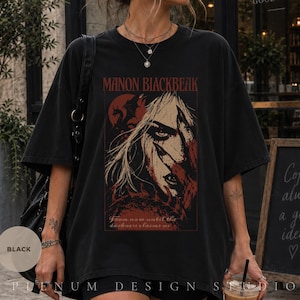 Puede incluir: Una camiseta negra de manga corta con un estampado gráfico de un personaje de pelo largo y la silueta de un dragón. El texto dice MANON BLACKBEAK y From now until the darkness claims us, utilizando tinta roja y blanca sobre la tela negra.