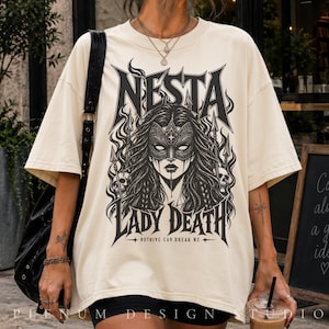 May include: An oversized cream-colored t-shirt featuring a black graphic print of a masked figure with long hair, surrounded by flames, spears, and skulls. The text reads NESTA LADY DEATH and NOTHING CAN BREAK ME in a bold, stylized gothic font.