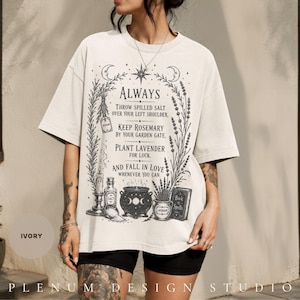 Puede incluir: Una camiseta de color marfil de manga corta con un estampado gráfico negro de elementos botánicos, un caldero, una vela y un libro de hechizos. El texto dice: Always throw spilled salt over your left shoulder, keep rosemary.