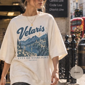 Könnte beinhalten: Ein elfenbeinfarbenes T-Shirt mit einem blauen Grafikdruck einer Bergstadtlandschaft. Der Text lautet "Velaris", "CITY OF STARLIGHT" und "PRYTHIAN". Das Design umfasst eine Mondsichel, Sterne und florale Elemente, mit Rundhalsausschnitt und kurzen Ärmeln.
