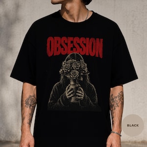 Puede incluir: Una camiseta negra de manga corta con un estampado gráfico de una persona sosteniendo un jarrón lleno de rosas frente a su rostro. La palabra OBSESSION está impresa en letras mayúsculas rojas grandes y desgastadas sobre la ilustración.