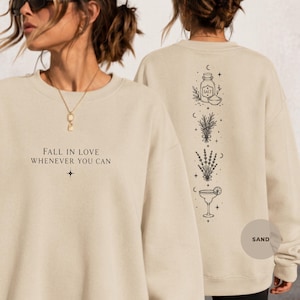 May include: A sand-colored crewneck sweatshirt featuring black line art. The front displays the text FALL IN LOVE WHENEVER YOU CAN with a star icon. The back shows a vertical design of a salt shaker, herbs, lavender, and a margarita glass.