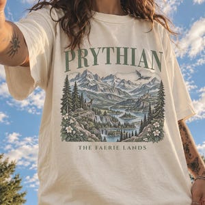 Może przedstawiać: Kremowy t-shirt z krótkim rękawem z nadrukiem graficznym zawierającym tekst PRYTHIAN oraz THE FAERIE LANDS. Ilustracja przedstawia górski krajobraz z sosnami, rzeką, jeleniem, zamkiem oraz latającym smokiem w stonowanych odcieniach.