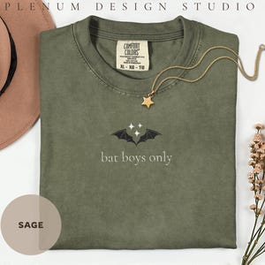 Może przedstawiać: Szałwiowo-zielony t-shirt z wyhaftowanym wzorem czarnych skrzydeł nietoperza i trzema małymi białymi gwiazdkami nad napisem "bat boys only" białą czcionką. Metka Comfort Colors przy kołnierzu wskazuje rozmiar XL-XG-TG.