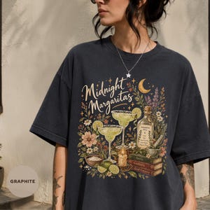 Puede incluir: Una camiseta color grafito con un estampado gráfico que incluye el texto Midnight Margaritas. El diseño muestra dos copas de margarita, una botella de poción, una pila de libros, una vela y varios elementos florales en tonos tierra.