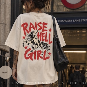 May include: An ivory short-sleeve t-shirt featuring a large graphic on the back. The design includes a black and white spotted horse rearing up, surrounded by red stars and the bold red text that reads RAISE HELL GIRL in a stylised font.