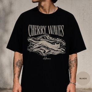 May include: A black short-sleeve t-shirt featuring a graphic print in a light beige color. The design displays the text "CHERRY WAVES" above an illustration of a cat swimming through stylized waves, with the word "deftones" printed below the cat.