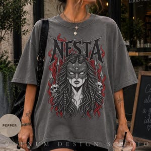 May include: A charcoal grey oversized t-shirt featuring a graphic print of a person wearing an ornate mask, surrounded by red flames, spears, and skulls. The text NESTA is printed in a bold, stylized black font above the central figure.