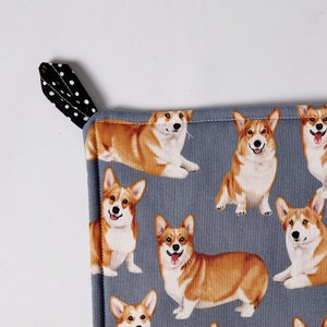May include: A gray and white pot holder with a pattern of brown and white corgis. The pot holder has a black and white polka dot hanging loop.