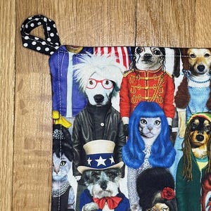 May include: Square, fabric pot holder with a colorful print of various pets dressed as famous people. The pot holder has a black and white polka dot loop for hanging. The text "PETS ROCK" is visible.