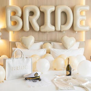 May include: A bridal suite decorated with ivory balloons spelling "BRIDE". A white tote bag with "BRIDE" embroidered on it, a captain's hat, and a satin robe are on the bed. Champagne and glasses are also present, creating a celebratory atmosphere.