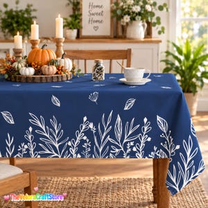 May include: A navy blue tablecloth featuring a white botanical print of leaves and stems along the bottom edge. The fabric is draped over a wooden table, which is set with a wooden tray holding three white candles and several small pumpkins.