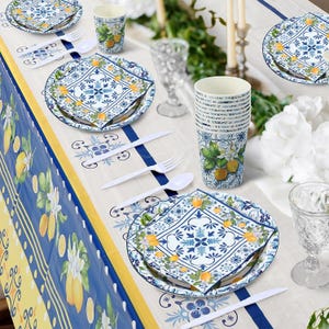 May include: A disposable party tableware set featuring a blue and white Mediterranean tile pattern with yellow lemon motifs. The set includes paper plates, napkins, and cups, accompanied by white plastic forks and spoons on a matching tablecloth.