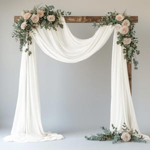 May include: A wooden arch frame draped with flowing white fabric, adorned with two floral arrangements featuring pale pink roses and green eucalyptus leaves. A small matching floral cluster rests on the floor at the base of the right side.