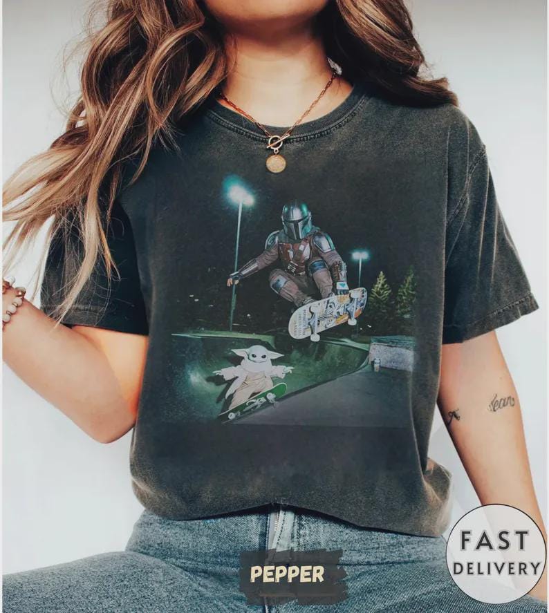 May include: Dark gray t-shirt featuring a graphic of The Mandalorian and Grogu skateboarding in a skate park. The Mandalorian is wearing his signature armor and Grogu is riding a skateboard. The shirt has a vintage, worn look.