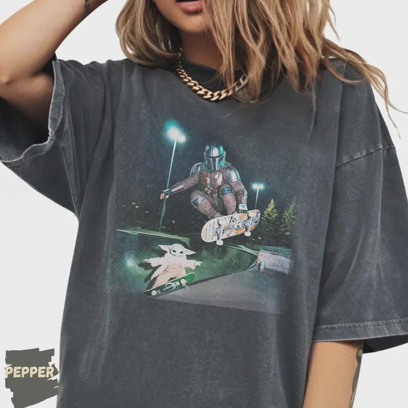 May include: Dark gray t-shirt featuring a graphic of the Mandalorian and Grogu skateboarding in a skate park. The design includes a detailed illustration of the characters and skateboards, set against a backdrop of a skate park with lights and trees.