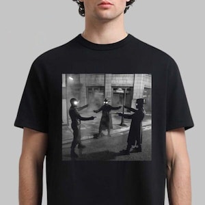 May include: Black t-shirt featuring a black and white image of three figures in a street scene, each pointing a gun. The figures are wearing dark clothing and hats, with bright lights on their heads. The background includes buildings and street signs.