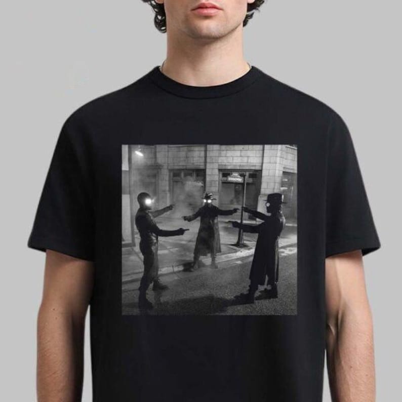 May include: Black t-shirt featuring a black and white image of three figures in a street scene, each pointing a gun. The figures are wearing dark clothing and hats, with bright lights on their heads. The background includes buildings and street signs.