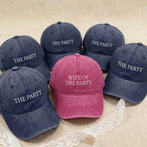 May include: A collection of baseball caps in navy blue and a single maroon cap. The navy caps have "THE PARTY" printed in white, while the maroon cap reads "WIFE OF THE PARTY". The caps are arranged on a neutral surface.