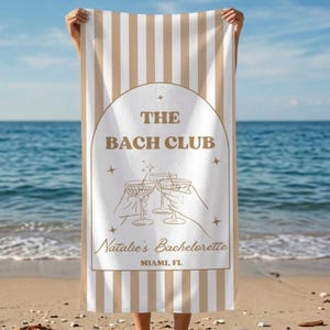 May include: A beach towel featuring vertical beige and white stripes. The center displays an arched frame with the text THE BACH CLUB, Natalie's Bachelorette, and MIAMI, FL, alongside a line-art illustration of hands holding cocktail glasses.