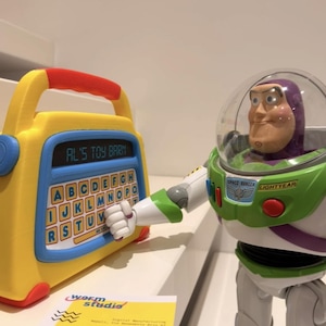 Könnte beinhalten: Ein gelbes und blaues Spielzeug mit dem Text "AL'S TOY BARN" und einer alphabetischen Tastatur wird von einer Buzz Lightyear-Actionfigur gehalten. Die Figur trägt einen grünen und weißen Raumanzug mit einem transparenten Helm. Eine Visitenkarte befindet sich im Vordergrund.