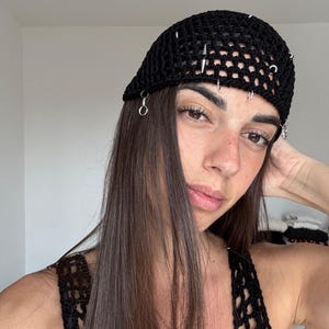 Op de afbeelding: Een zwarte gehaakte hoed met zilveren piercings en ringen. De hoed heeft een netachtig ontwerp en wordt op het hoofd gedragen. De hoed is een modeaccessoire.