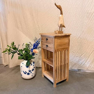 Puede incluir: Una mesa auxiliar de madera clara con dos cajones y laterales de listones. Una figura de pato de madera se encuentra en la parte superior. Un jarrón blanco con diseños florales azules contiene un ramo de flores.