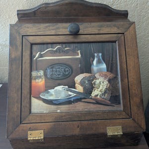 Può includere: Un portapane in legno marrone scuro con uno sportello frontale inclinato dotato di un pannello in vetro. Il vetro mostra un'immagine stampata di una pagnotta, un barattolo di marmellata, una bottiglia di latte e un burriera con coltello. Il contenitore ha un pomello rotondo e cerniere in ottone.