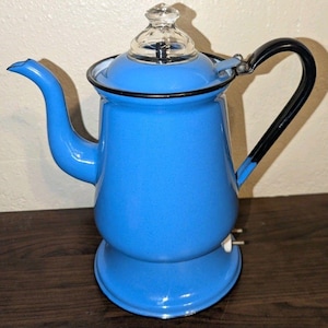 May include: A bright blue enamel coffee percolator with a black handle and a clear glass knob on the lid. The pot features a curved spout, a wide base, and a black rim around the top edge. A small white electrical plug is visible at the bottom.