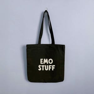 May include: Black tote bag with the words "EMO STUFF" printed in white. The bag is hanging against a light blue wall. The tote bag has long handles and is made of a sturdy material.