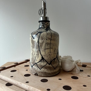 May include: A ceramic oil dispenser with a cream-colored glaze, featuring black hand-painted line patterns and dark speckles. The bottle is topped with a silver-colored metal pour spout with a hinged lid, designed for controlled liquid dispensing.