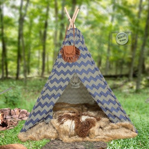 Puede incluir: Una tienda de campaña tipi con estampado de chevron azul y beige con una alfombra de piel sintética en el interior. El tipi está instalado en un entorno de bosque con una pequeña fogata delante.