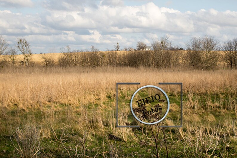 May include: A field of tall, dry grass with a few trees in the background. The sky is cloudy and blue. A watermark in the foreground reads "Eliejo Fotos".