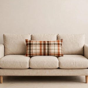 May include: A rectangular throw pillow featuring a plaid pattern in shades of brown, orange, and cream. The pillow is positioned horizontally on the center seat cushion of a solid light beige sofa with three back cushions and a smooth fabric finish.