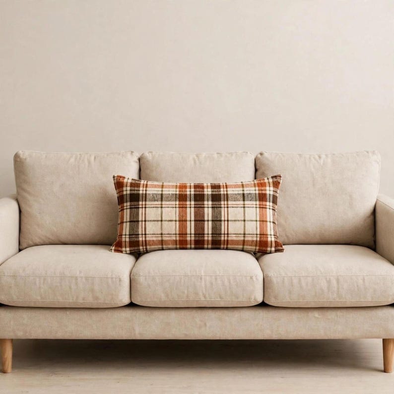 May include: A rectangular throw pillow featuring a plaid pattern in shades of brown, orange, and cream. The pillow is positioned horizontally on the center seat cushion of a solid light beige sofa with three back cushions and a smooth fabric finish.