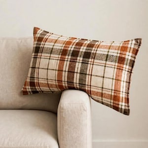 May include: A rectangular throw pillow featuring a plaid pattern in shades of cream, burnt orange, dark brown, and olive green. The fabric has a soft, textured appearance with a classic intersecting grid design across the entire surface.